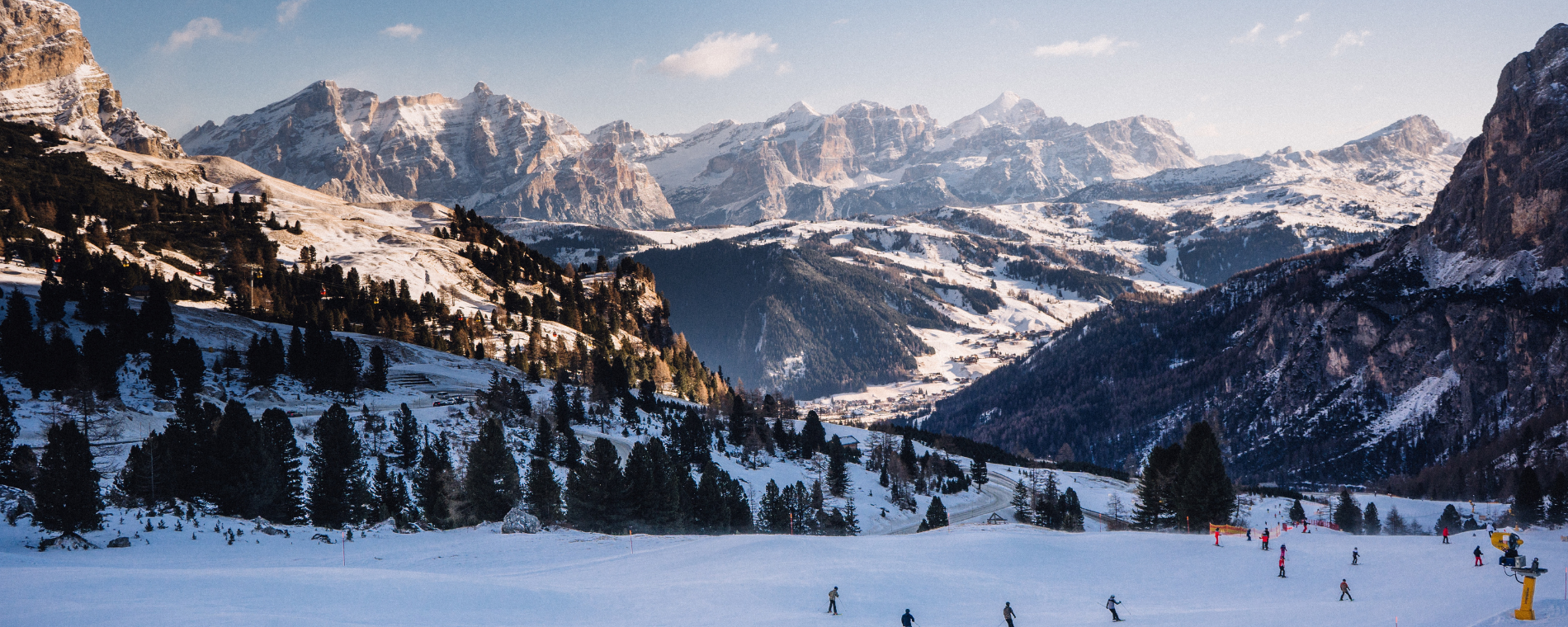 Vast landscape with blue sky and snowy mountains in the background. Skiers skiing on slope in foreground.