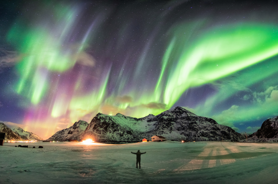 travel trends - noctourism. Person standing in front of a snowy mountain with the Northern Lights above them.